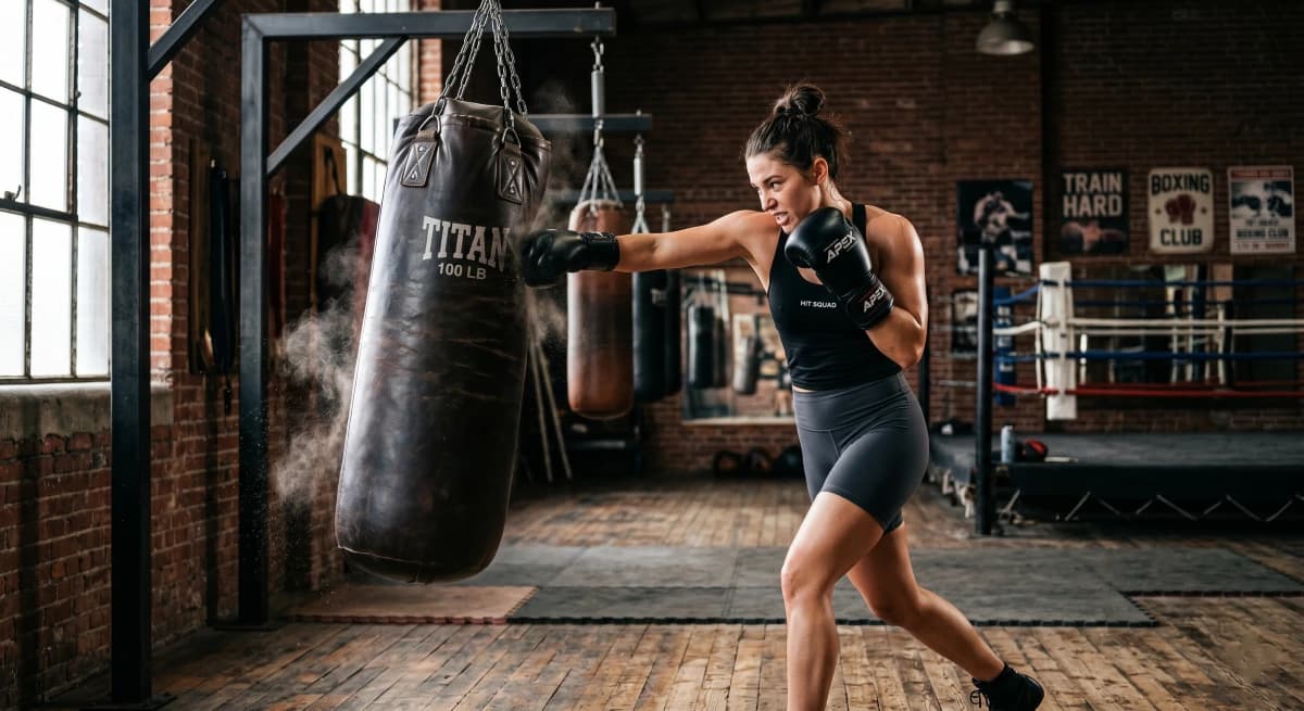 A female boxer hitting a heavy bag with proper stance and boxing gloves during training in a gym.
